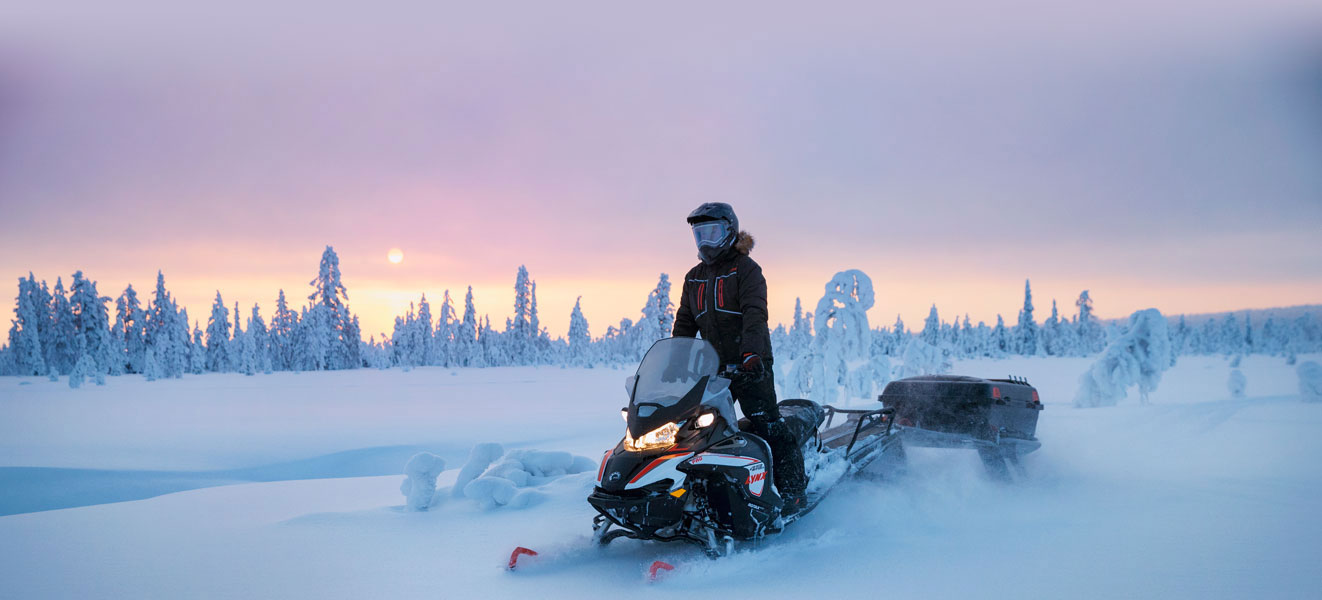 A man is riding his Lynx 49 Ranger Snowmobile Model on a snowy road at sunset