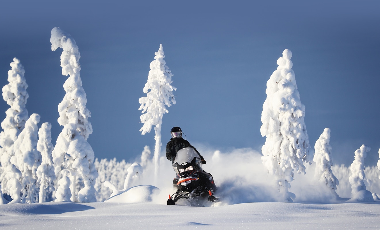 A man is riding his Lynx Commander Snowmobile Model throught the snowy forest under the sun