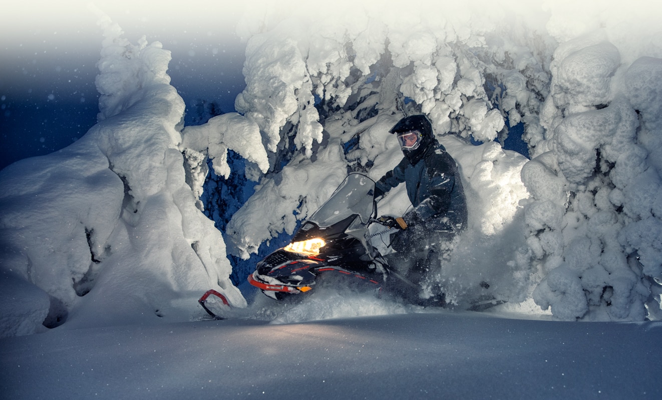 A man is driving his Lynx Commander Snowmobile Model throught the snowy forest during the night