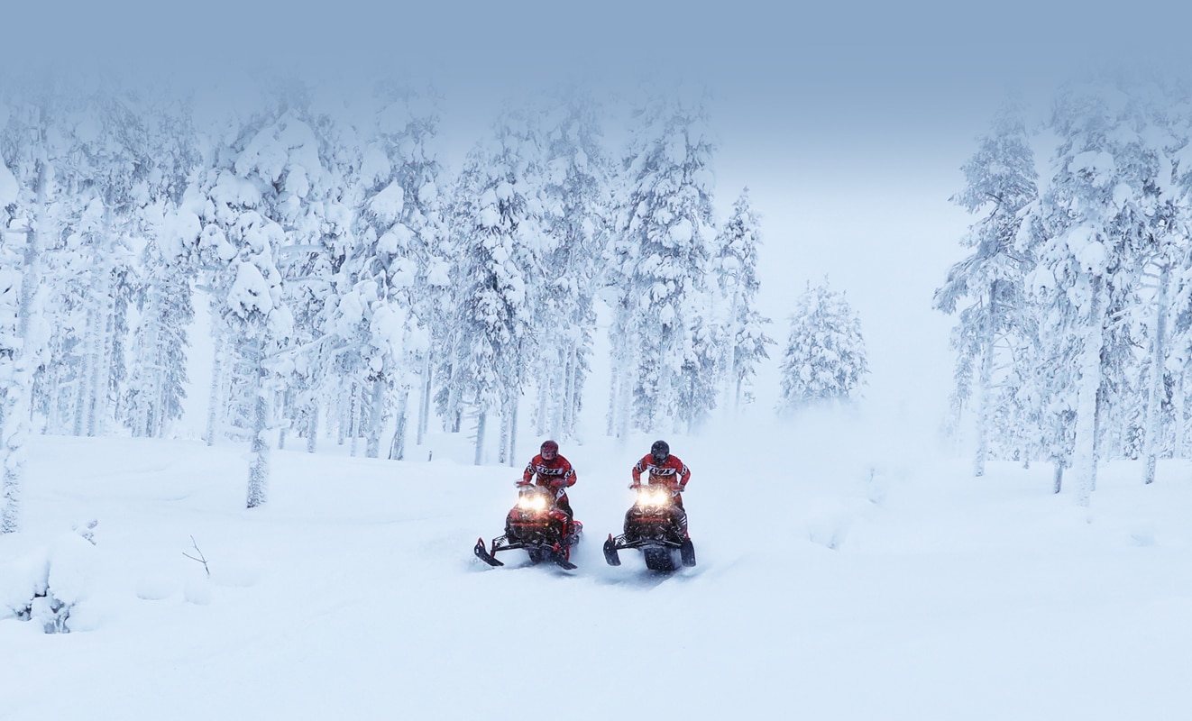 Dvojica muškaraca voze se snježnom cestom kroz šumu sa svojim modelom motornih sanki Lynx Rave Re