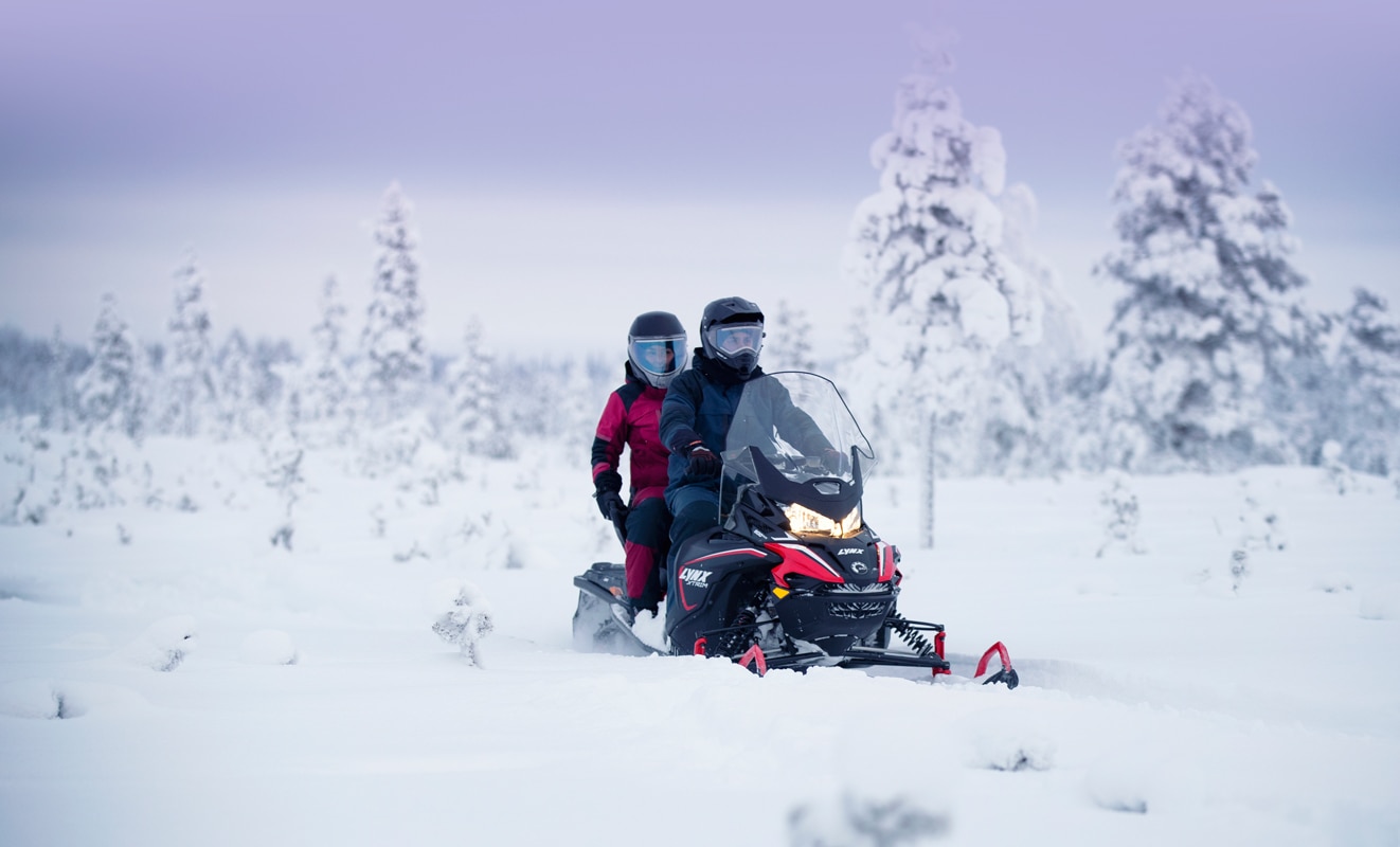 A couple is riding a Lynx Xtrim Snowmobile Model on a snowy road