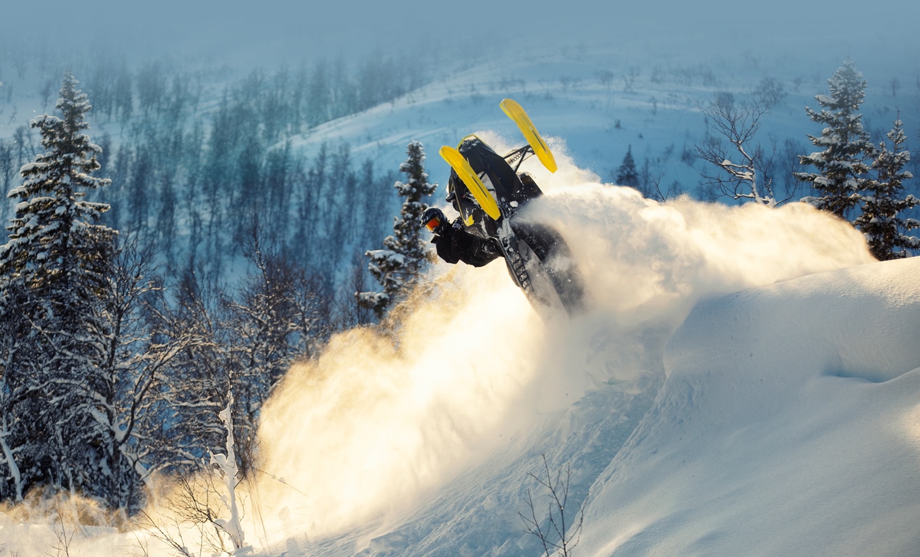 A man is doing a backflip with his Lynx Xtrim Snowmobile Model on the top of a snowy hill
