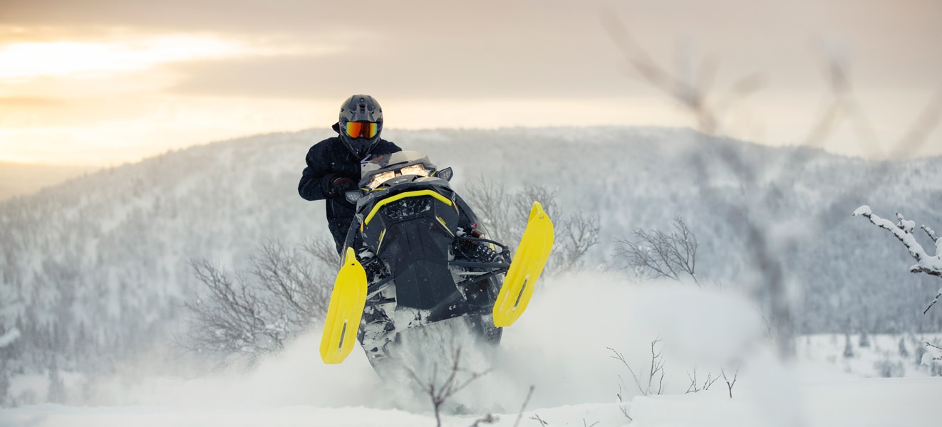 A man is jumping with his Lynx Xtrim Snowmobile Model on the top of a snowy hill