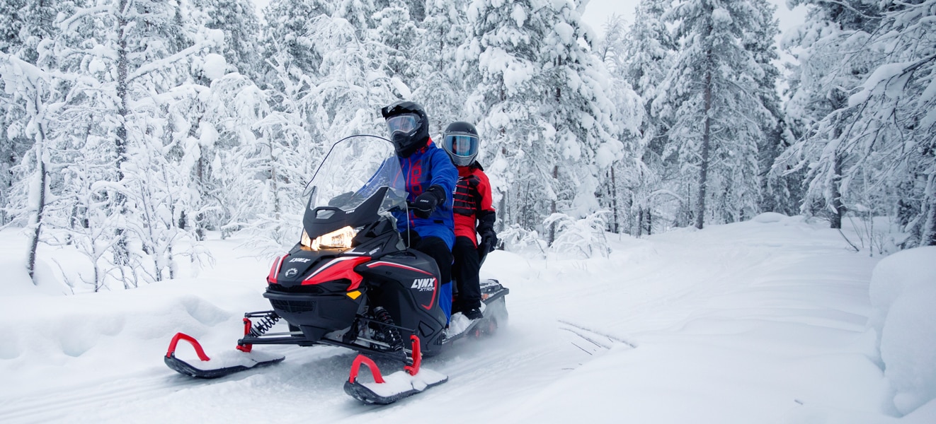 A couple is riding in the middle of a snowy forest on their Lynx Xtrim Snowmobile Model 