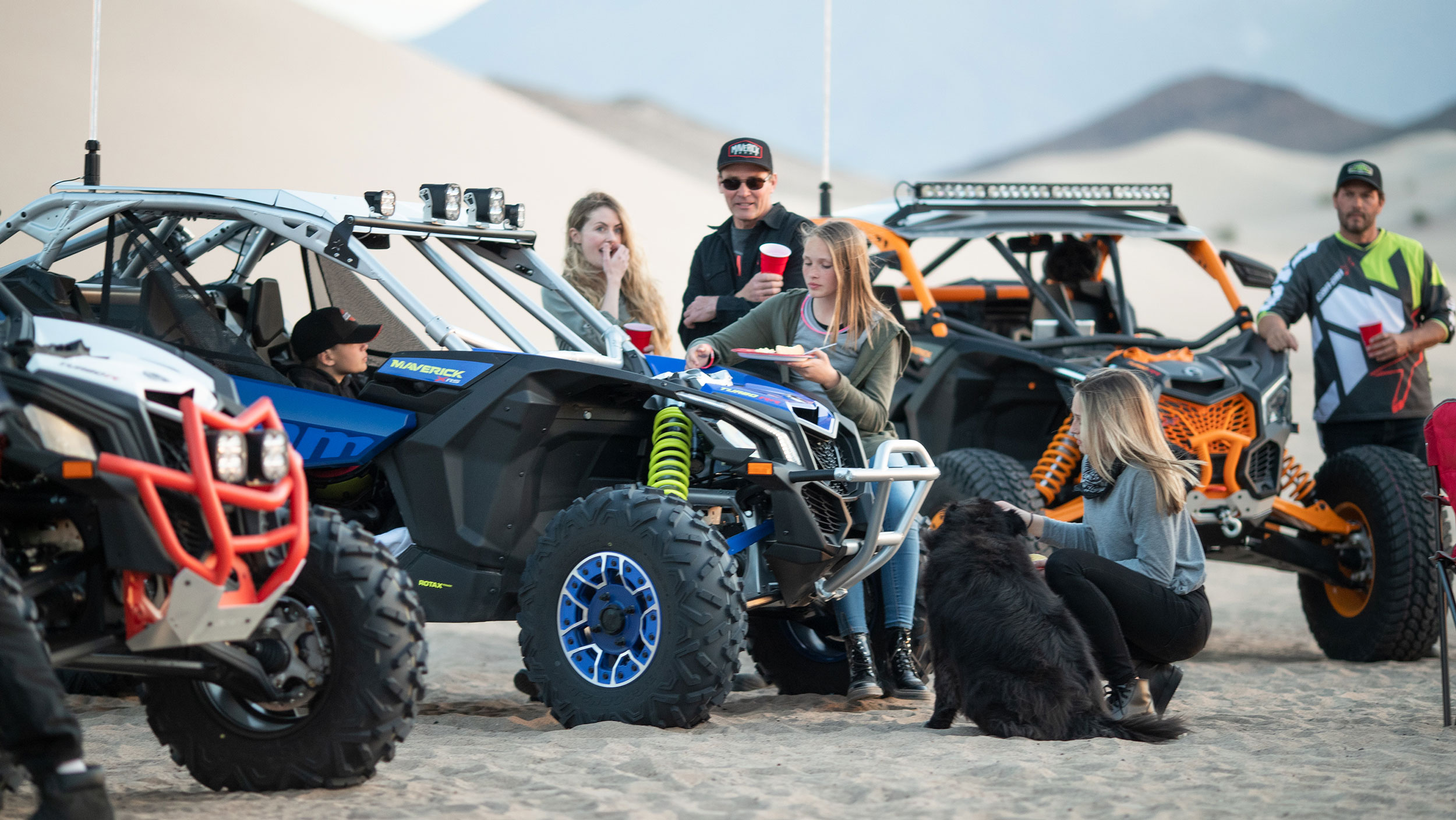 egy csapat fiatal bandázik a homokos terepen három maverick mellett és egy kutyával